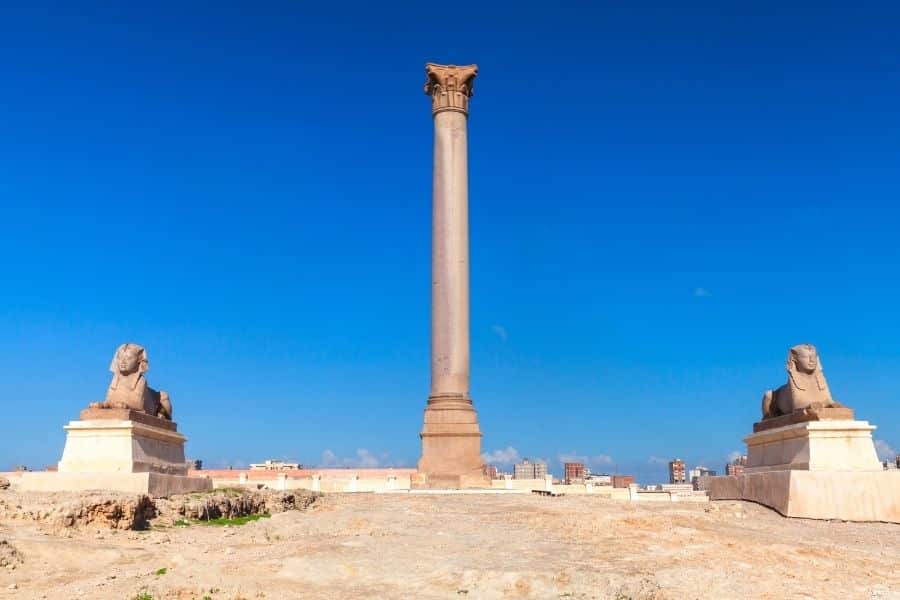 Pompey's Pillar in Alexandria, Egypt, flanked by two ancient sphinx statues, standing against a clear blue sky.