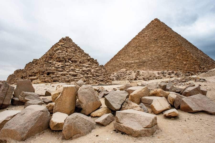 The iconic pyramids on the Giza Plateau, showcasing weathered stone structures under a cloudy sky, surrounded by scattered rocks.