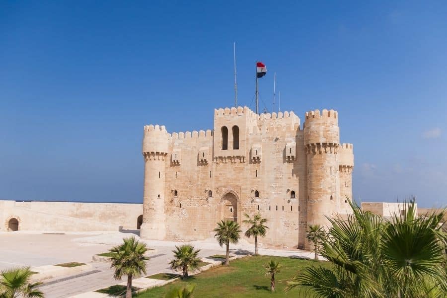 Exterior view of Qaitbay Fort in Alexandria, Egypt, with tall stone walls, towers, and an Egyptian flag on top.
