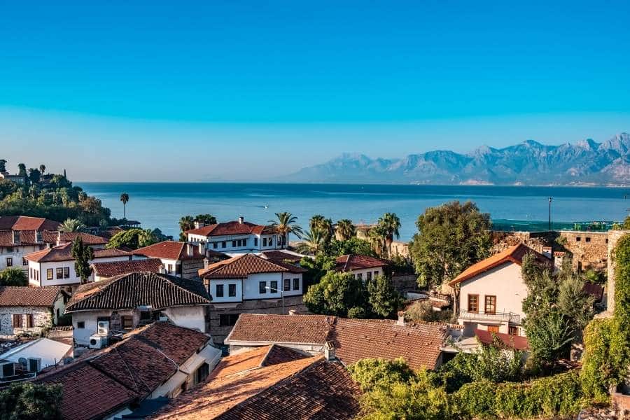 Rooftop view of Kaleiçi, Antalya, featuring traditional red-tiled houses, lush greenery, and the Mediterranean Sea with distant mountains.