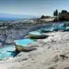 View of the terraced thermal pools of Hierapolis-Pamukkale in Turkey, showcasing bright white limestone formations filled with mineral-rich turquoise waters under a clear blue sky, with ancient Greco-Roman ruins visible in the background.