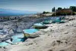 View of the terraced thermal pools of Hierapolis-Pamukkale in Turkey, showcasing bright white limestone formations filled with mineral-rich turquoise waters under a clear blue sky, with ancient Greco-Roman ruins visible in the background.