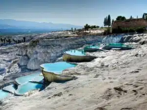 View of the terraced thermal pools of Hierapolis-Pamukkale in Turkey, showcasing bright white limestone formations filled with mineral-rich turquoise waters under a clear blue sky, with ancient Greco-Roman ruins visible in the background.