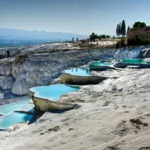 View of the terraced thermal pools of Hierapolis-Pamukkale in Turkey, showcasing bright white limestone formations filled with mineral-rich turquoise waters under a clear blue sky, with ancient Greco-Roman ruins visible in the background.
