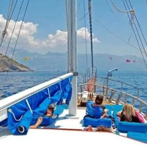 A group of people enjoying a sunny day on the deck of a sailboat with a clear view of the sea and mountains in the distance.