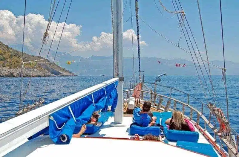 A group of people enjoying a sunny day on the deck of a sailboat with a clear view of the sea and mountains in the distance.