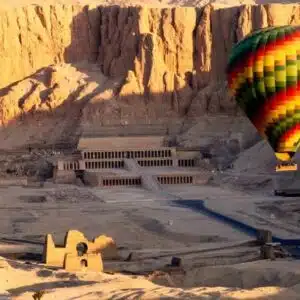 Colorful hot air balloon floats over the Temple of Hatshepsut in Luxor, Egypt, with sunlit cliffs in the background.