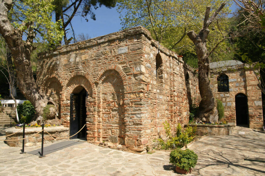 Stone-brick House of the Virgin Mary in Ephesus surrounded by trees and flowers under clear blue skies.