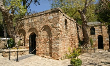 Stone-brick House of the Virgin Mary in Ephesus surrounded by trees and flowers under clear blue skies.