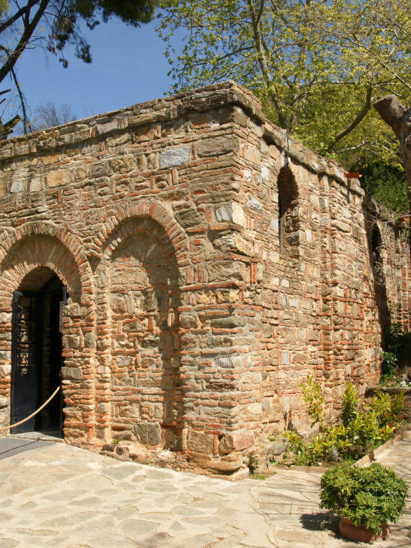 Stone-brick House of the Virgin Mary in Ephesus surrounded by trees and flowers under clear blue skies.