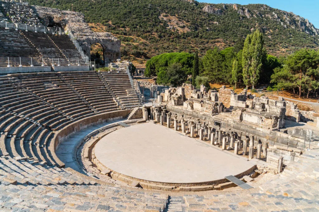 Wide-angle view of the Grand Theatre of Ephesus with stone seating, stage ruins, and forested hills in the background.