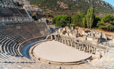 Wide-angle view of the Grand Theatre of Ephesus with stone seating, stage ruins, and forested hills in the background.