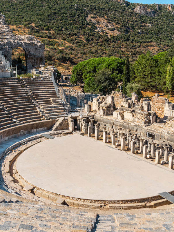 Wide-angle view of the Grand Theatre of Ephesus with stone seating, stage ruins, and forested hills in the background.