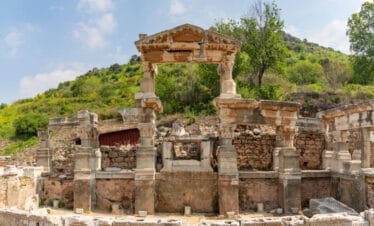 Ruins of the Fountain of Trajan in Ephesus with preserved Roman columns and stone reliefs amid lush hillside.