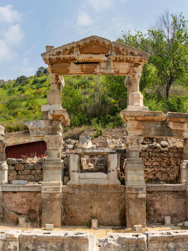 Ruins of the Fountain of Trajan in Ephesus with preserved Roman columns and stone reliefs amid lush hillside.