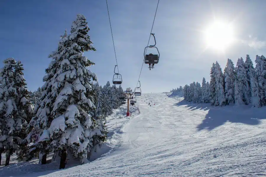 A ski lift with empty chairs ascends over a snowy slope dotted with pine trees under a bright blue sky.