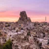Uçhisar Castle rises above stone houses and cave dwellings under a colorful sunset sky in Cappadocia, Turkey.