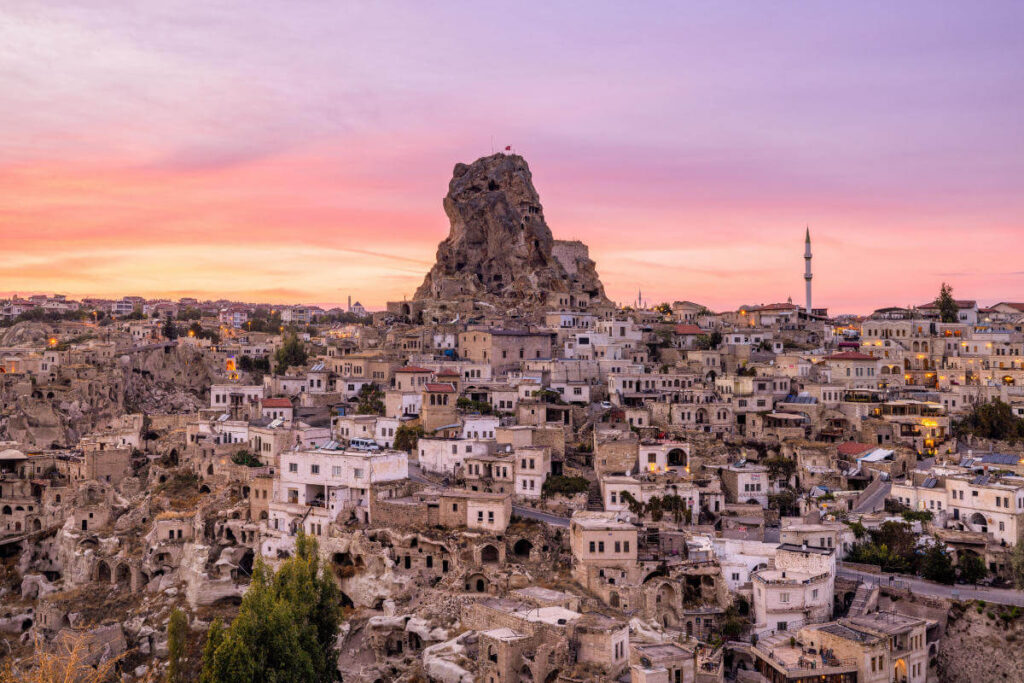 Uçhisar Castle rises above stone houses and cave dwellings under a colorful sunset sky in Cappadocia, Turkey.