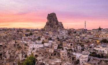 Uçhisar Castle rises above stone houses and cave dwellings under a colorful sunset sky in Cappadocia, Turkey.