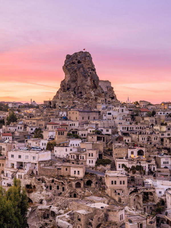 Uçhisar Castle rises above stone houses and cave dwellings under a colorful sunset sky in Cappadocia, Turkey.