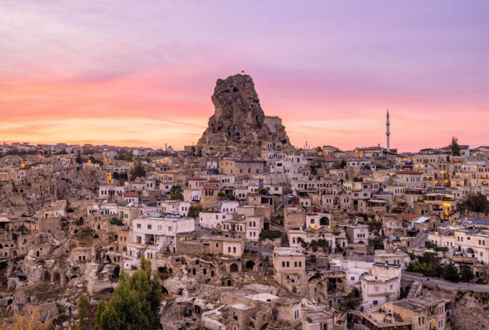 Uçhisar Castle rises above stone houses and cave dwellings under a colorful sunset sky in Cappadocia, Turkey.