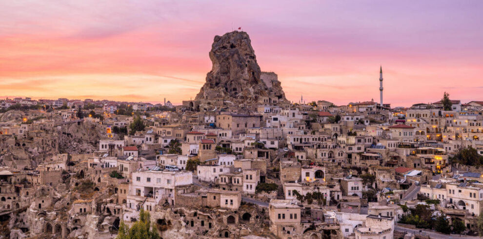 Uçhisar Castle rises above stone houses and cave dwellings under a colorful sunset sky in Cappadocia, Turkey.