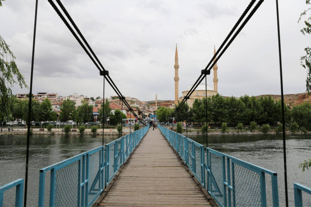 Blue pedestrian suspension bridge over river leading to mosque with twin minarets in Avanos, Cappadocia.