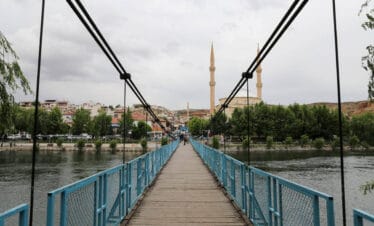 Blue pedestrian suspension bridge over river leading to mosque with twin minarets in Avanos, Cappadocia.