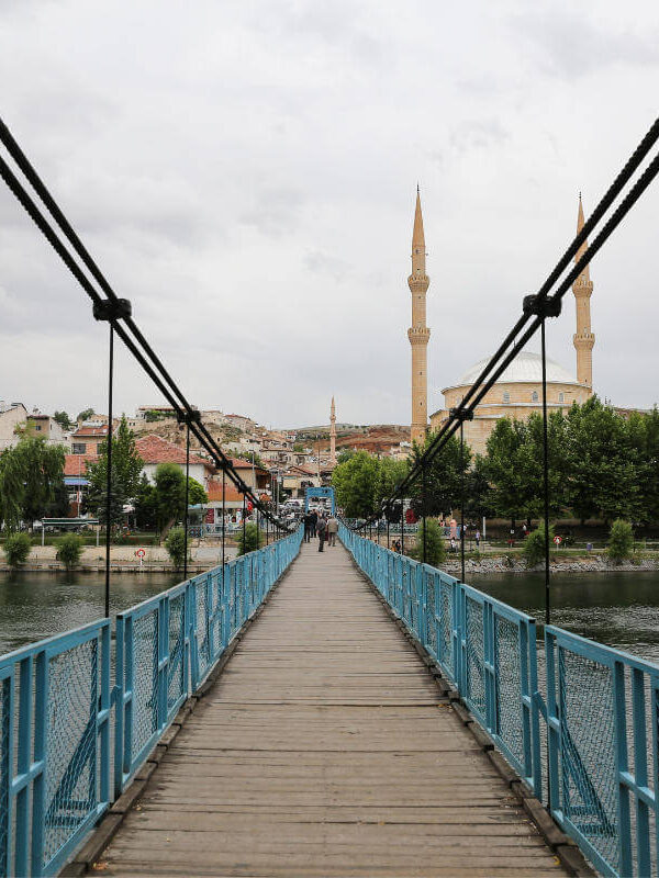 Blue pedestrian suspension bridge over river leading to mosque with twin minarets in Avanos, Cappadocia.