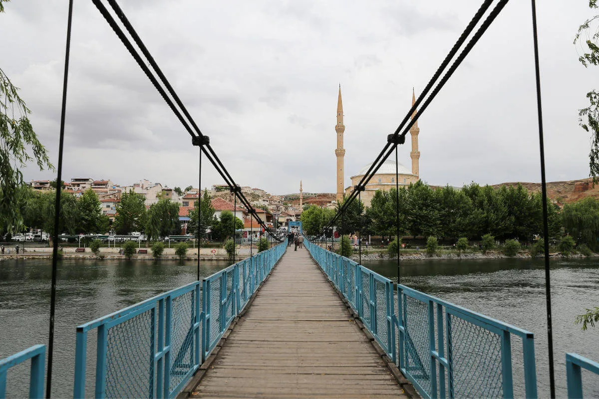 Blue pedestrian suspension bridge over river leading to mosque with twin minarets in Avanos, Cappadocia.