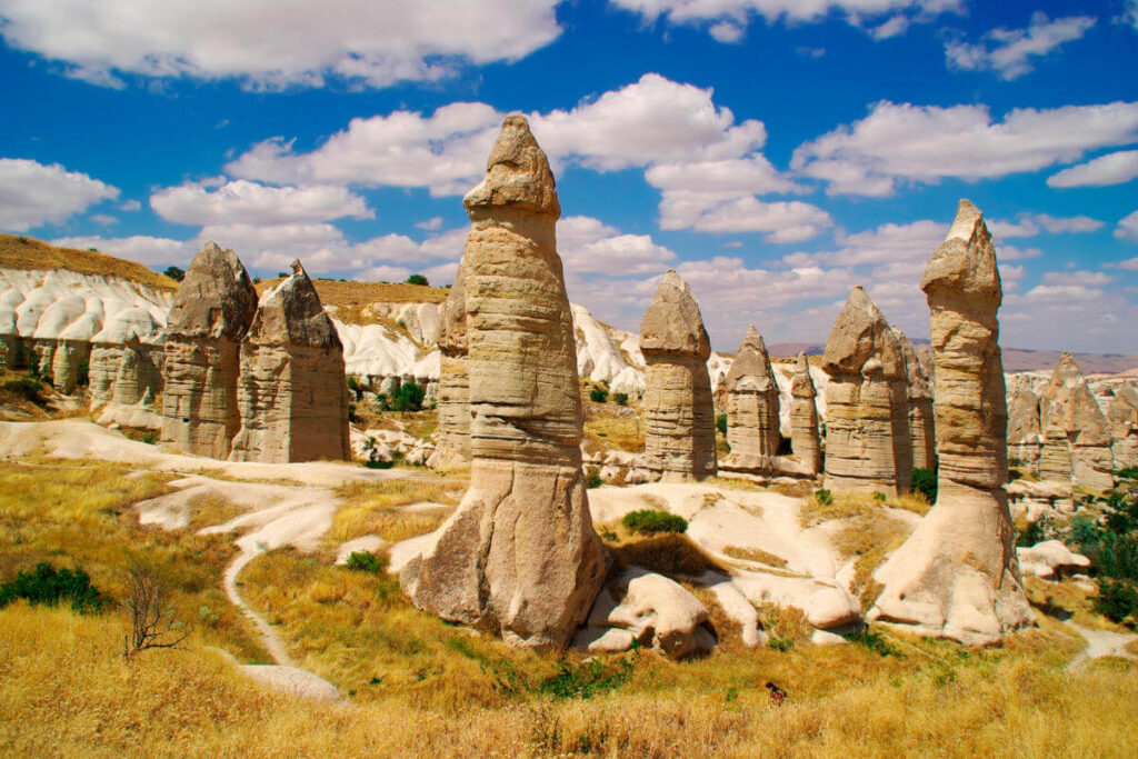 Tall, phallic-shaped fairy chimneys in Love Valley, Cappadocia, under a bright blue sky with scattered clouds.