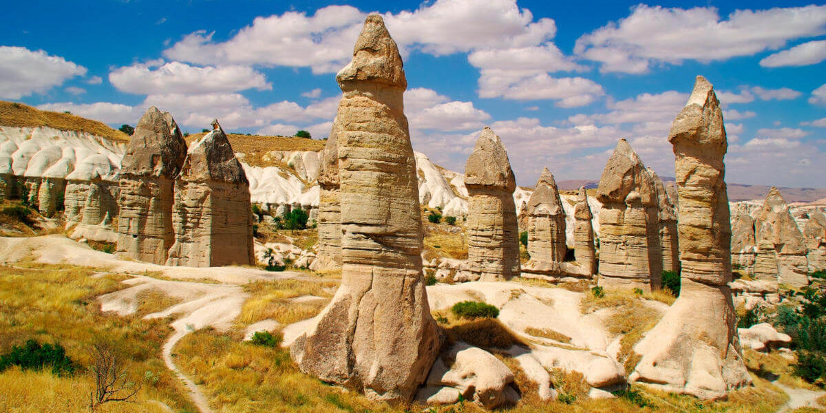 Tall, phallic-shaped fairy chimneys in Love Valley, Cappadocia, under a bright blue sky with scattered clouds.