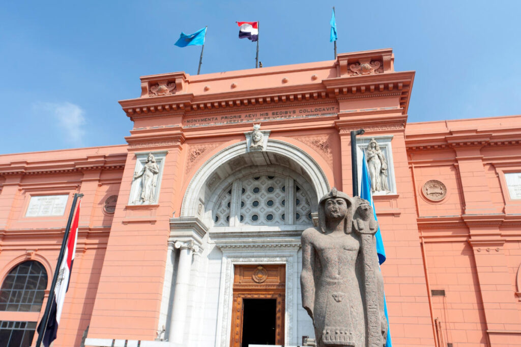 Grand facade of the Egyptian Museum in Cairo with ancient pharaoh statue and flags under clear blue sky.