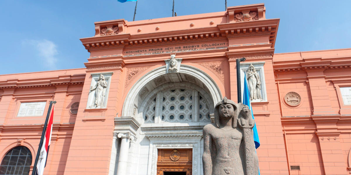 Grand facade of the Egyptian Museum in Cairo with ancient pharaoh statue and flags under clear blue sky.