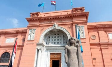 Grand facade of the Egyptian Museum in Cairo with ancient pharaoh statue and flags under clear blue sky.