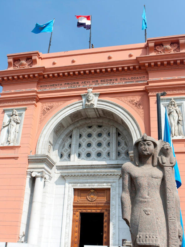 Grand facade of the Egyptian Museum in Cairo with ancient pharaoh statue and flags under clear blue sky.