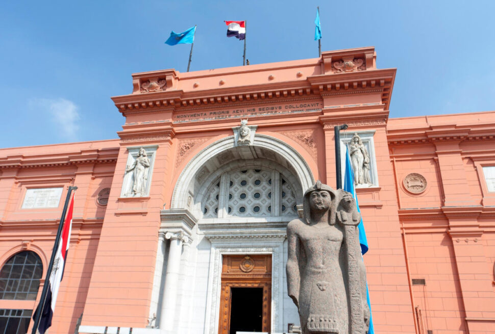 Grand facade of the Egyptian Museum in Cairo with ancient pharaoh statue and flags under clear blue sky.