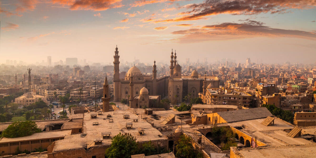 Skyline of Cairo, Egypt at sunrise with historic mosques and minarets surrounded by dense urban architecture and colorful clouds.