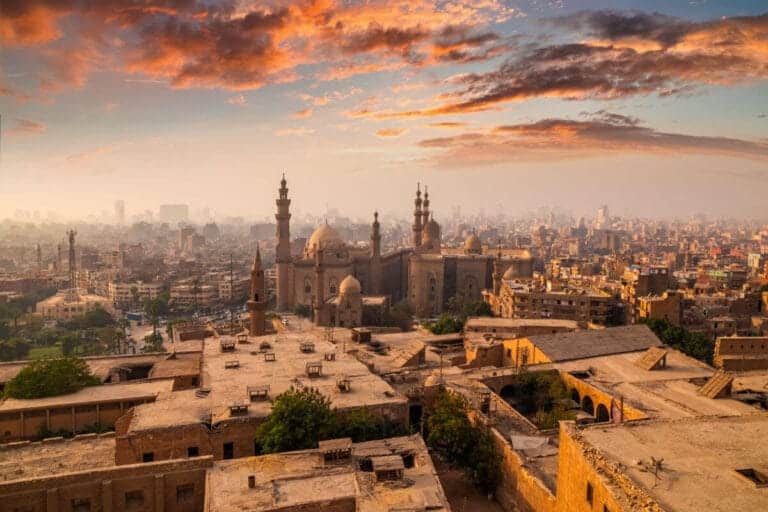 Skyline of Cairo, Egypt at sunrise with historic mosques and minarets surrounded by dense urban architecture and colorful clouds.