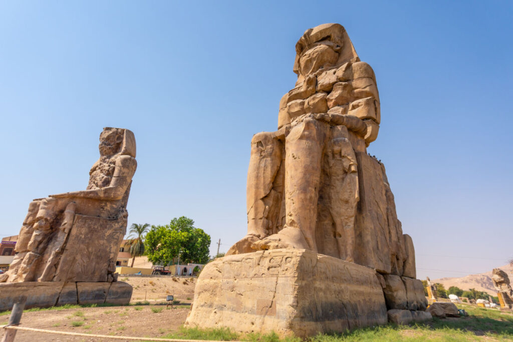 Colossi of Memnon statues in Luxor, Egypt, showing two massive seated stone figures under a clear blue sky.