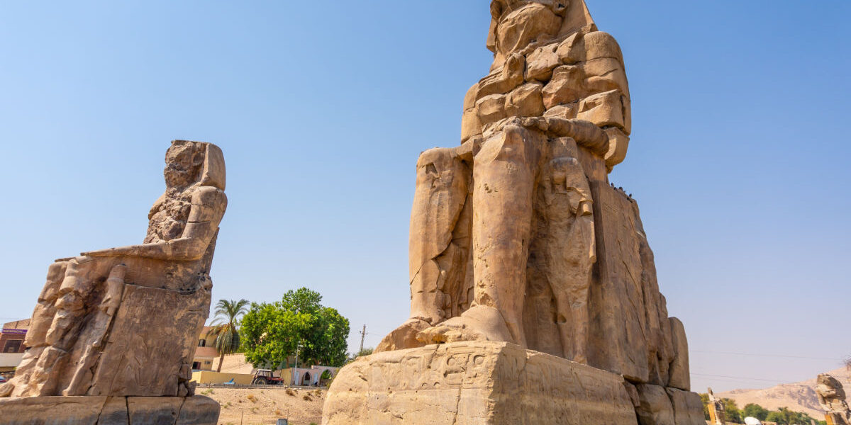 Colossi of Memnon statues in Luxor, Egypt, showing two massive seated stone figures under a clear blue sky.