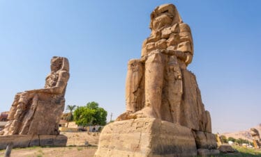 Colossi of Memnon statues in Luxor, Egypt, showing two massive seated stone figures under a clear blue sky.