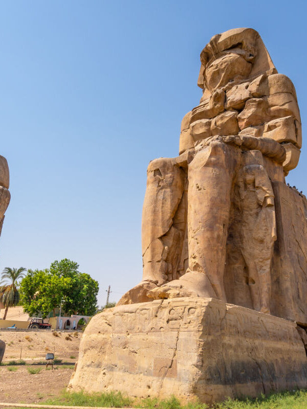 Colossi of Memnon statues in Luxor, Egypt, showing two massive seated stone figures under a clear blue sky.