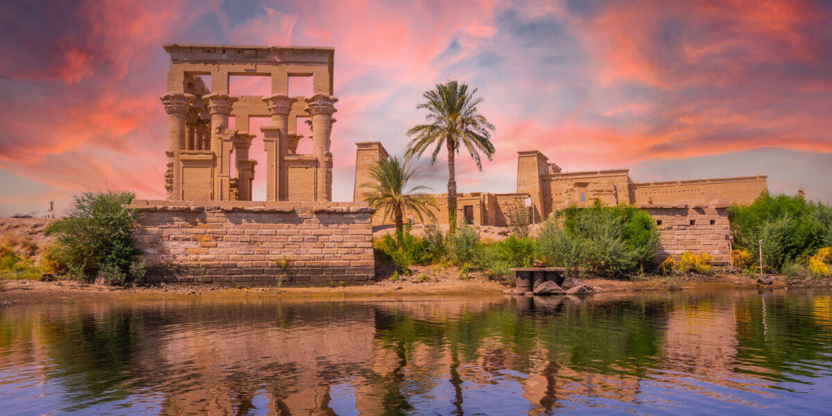 Philae Temple in Aswan, Egypt, reflected in the Nile River at sunset with vivid pink and orange skies and lush greenery.