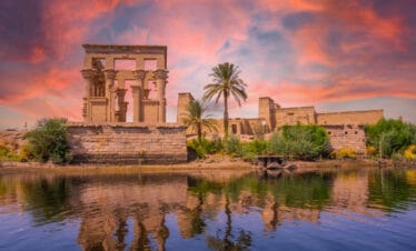 Philae Temple in Aswan, Egypt, reflected in the Nile River at sunset with vivid pink and orange skies and lush greenery.