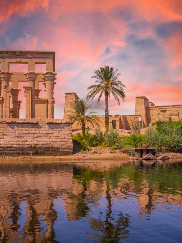 Philae Temple in Aswan, Egypt, reflected in the Nile River at sunset with vivid pink and orange skies and lush greenery.