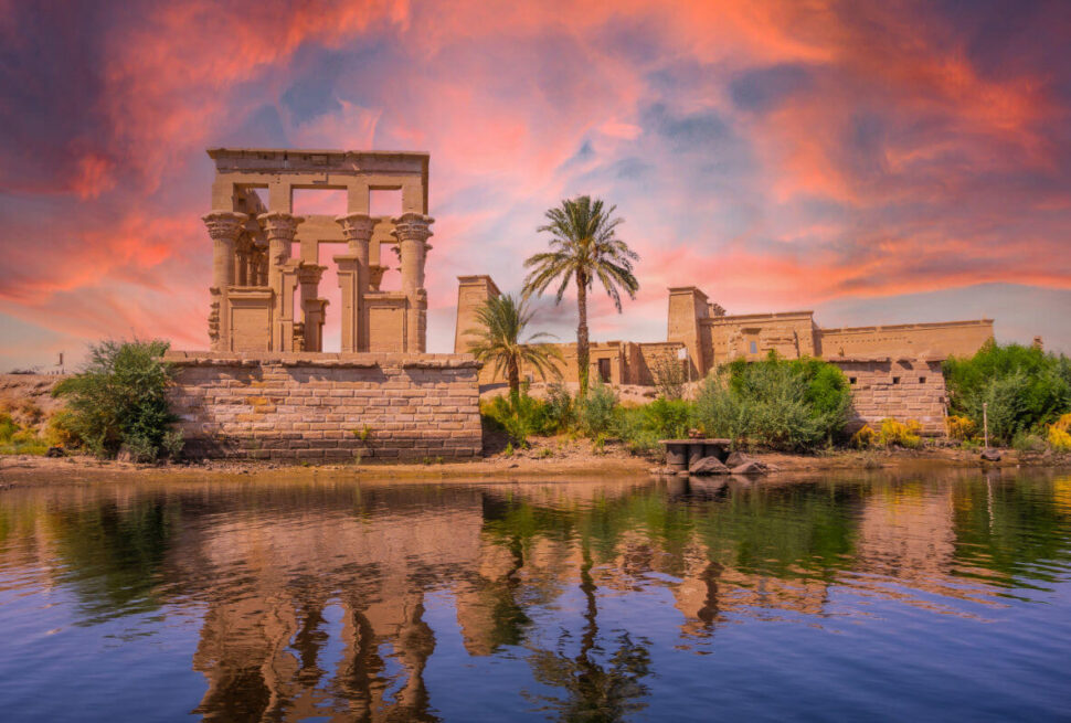 Philae Temple in Aswan, Egypt, reflected in the Nile River at sunset with vivid pink and orange skies and lush greenery.