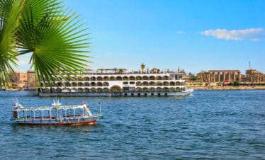 Cruise ship and small boat sailing on the Nile River in Luxor, Egypt, with ancient temple ruins and palm leaves in the background.