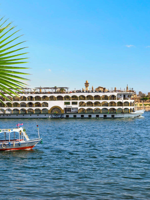 Cruise ship and small boat sailing on the Nile River in Luxor, Egypt, with ancient temple ruins and palm leaves in the background.