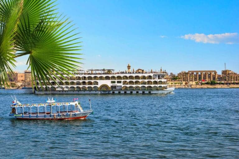 Cruise ship and small boat sailing on the Nile River in Luxor, Egypt, with ancient temple ruins and palm leaves in the background.